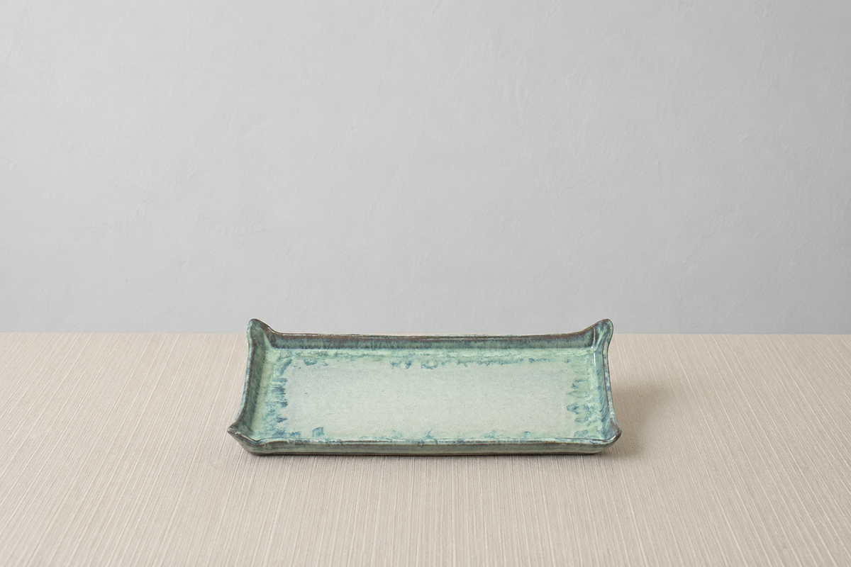 A rectangular ceramic serving tray with a glossy, textured glaze in shades of light blue-green and teal, set against a neutral light gray background and a beige textured tabletop.